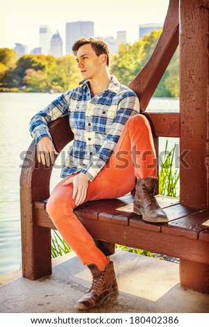 Young Man Relaxing Outside. Dressing in a long sleeve shirt, red pants, brown boots, a young guy is causally sitting on the bench by a lake. City buildings in background. Instagram Hefe filter effect.