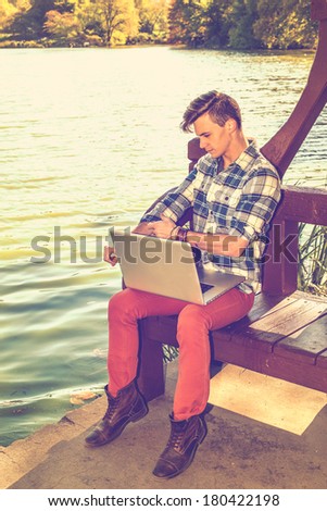 Dressing in a long sleeve, patterned shirt, red pants, brown boots, rolling over sleeves, one young guy is working on a laptop computer by a lake in afternoon. / Student Studying Outside