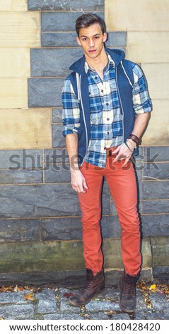 Dressing in a sleeveless hoodie, a long sleeve, patterned shirt, red pants, brown boots, one young man is standing by a modern fashion style wall, looking at you. / Fashion Boy