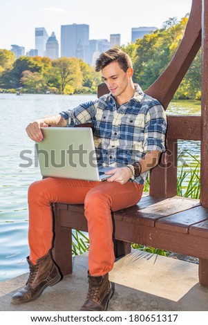 Dressing in a long sleeve, patterned shirt, red pants, brown boots, one young man is working on a laptop computer by a lake in afternoon. Handsome fashion guy reading outside