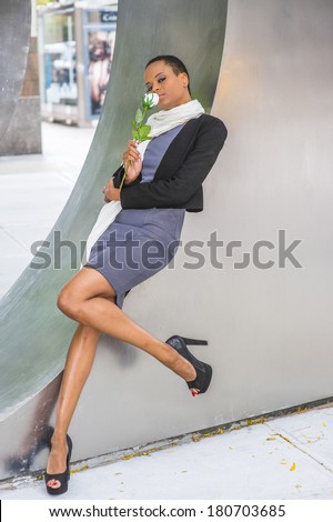 Pretty Black Woman with White Flower. Dressing in woolen cropped jacket, dress, long scarf, high heels, holding white rose,  a young black fashion lady is relying on structure, smelling, thinking.