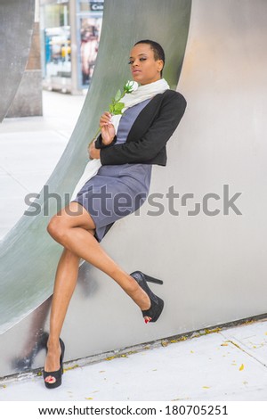 Pretty Black Woman with White Flower. Dressing in woolen cropped jacket, dress, long scarf, high heels, holding white rose,  a young black fashion lady is relying on a metal structure, thinking.
