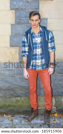 Fashion Boy. Dressing in a sleeveless hoodie, a long sleeve, patterned shirt, red pants, brown boots, one young man is standing by a modern fashion style wall, looking at you.