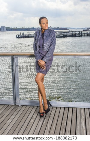 Black Woman Thinking Outside. Dressing in a gray patterned faux fur jacket,  a woolen fitted dress and open toes shoes, a young professional lady is standing on the dock by a river, waiting for you.