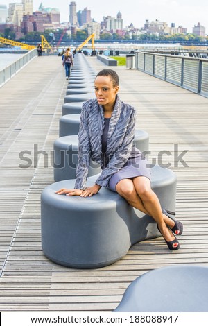 Black Woman Thinking Outside. Dressing in a gray patterned faux fur jacket, a woolen fitted dress, open toes shoes, a young professional lady is sitting on a modern style bench, relaxing, thinking.