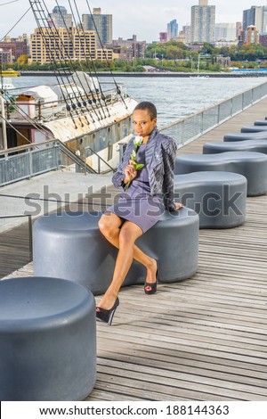 Dressing in faux fur jacket, a woolen fitted dress, open toes shoes, holding a white flower,  a young black woman is sitting on a deck, thinking about you. The background is a harbor.