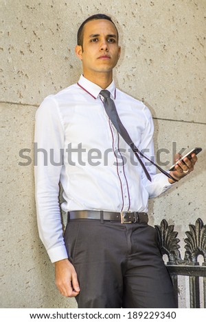 Man Thinking and Texting Outside.  Dressing in a white shirt, a black tie, holding a cell phone, a young handsome guy is leaning against the wall outside, into deeply thinking.