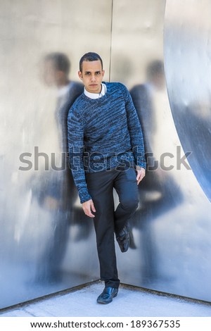 Man Casual Fashion.  Dressing in a blue patterned sweater, black pants, leather shoe, a young attractive guy is standing by a metal structure, waiting for you.