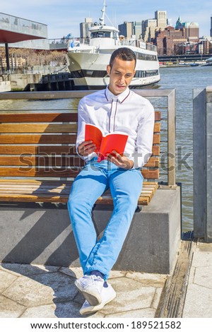 Man Reading Outside. Wearing a white shirt, blue pants, white sneakers, a young handsome student is sitting on a chair under sunshine in a harbor, reading a red book.