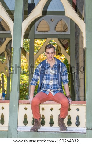 Young Man Relaxing Outside. Dressing in a blue and white pattern shirt,  a blue hood vest,  red jeans and brown leather boot shoes, a young guy is sitting by a pavilion, waiting for you.