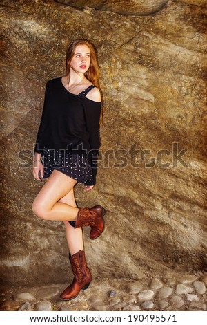 Fashion Girl. Dressing in black sweater, showing a shoulder,  patterned skirt, brown boots,  a teenage girl is standing by rocks, bending a leg, confidently looking forward. Instagram Hefe effect.