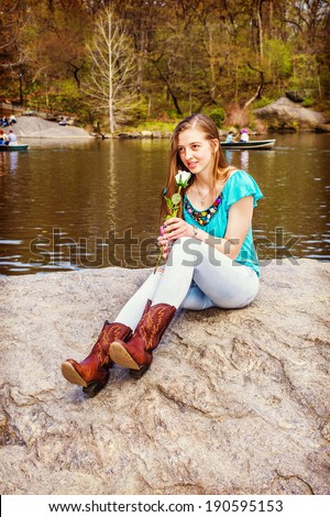 Missing You. Dressing in a blue sleeveless top, fashionable jeans, brown boots, a blonde teenage girl is sitting on rocks by a lake, smelling a white flower, thinking. Instagram Hefe effect.