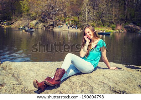 Calling on Park. Dressing in a blue sleeveless top, fashionable jeans, brown boots, a blonde teenage girl is sitting on rocks by a lake, talking on her cell phone. Instagram X-Pro II effect.