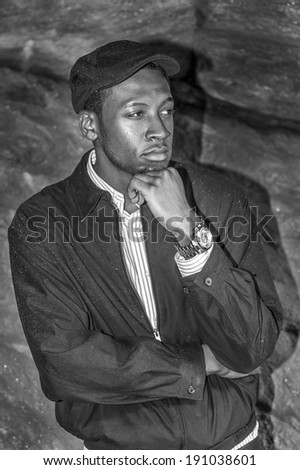 Rainy Day, a little foggy, wet feel. Wearing a waterproof jacket, a ivy cap,  a young handsome black man is standing by rocks, one hand touching his chin into deeply thinking. Clothes is wet by rain.