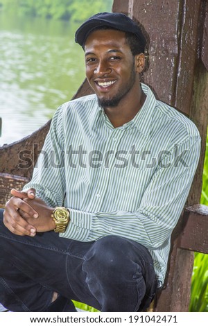 Rainy Day, a little foggy, wet feel. Dressing in a long sleeves shirt, a ivy cap,  a young handsome black guy is squatting by a lake in a raining, foggy day, smilingly looking at you. Man relaxing.