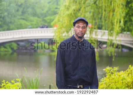 In the rain, a foggy, wet , blur feel. Wearing a waterproof jacket, a ivy cap,  a young handsome black man is standing in the rain on the park, seriously looking at you.