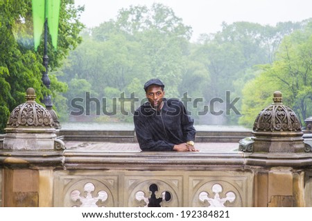 In the rain, a foggy, wet , blur feel. Wearing a waterproof jacket, a ivy cap,  a young black guy is standing by a fence, patiently waiting for you to meet. Man waiting in the rain.