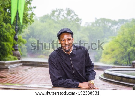 In the rain, a foggy, wet , blur feel. Wearing a waterproof jacket, a ivy cap,  a young black guy is standing in the rain, smiling, looking at you. Man waiting for you in the rain.
