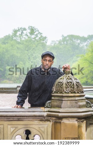 In the rain, a foggy, wet , blur feel. Wearing a waterproof jacket, a ivy cap,  a young black guy is standing by a fence, patiently waiting for you to meet. Man waiting in the rain.