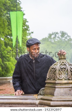 In the rain, a foggy, wet , blur feel. Wearing a waterproof jacket, a ivy cap,  a young black man is standing outside in the rain, sad, depressed, into deeply thinking.