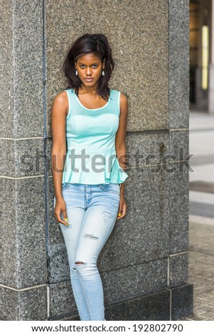 Portrait of Young Girl. Wearing a green tank top, fashionable jeans, drop earrings, a young pretty black woman is standing against the wall, lowering her forehead, intensely looking at you.