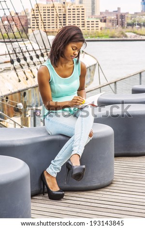 Girl Reading Outside. Wearing a green tank top, blue fashionable jeans, a young pretty woman is sitting on the deck in a harbor, crossing legs, looking down, reading a book on her laptop,