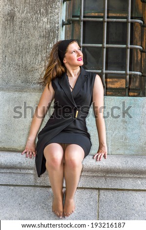 Sexy Woman Sitting Outside. Wearing a black sleeveless trench coat dress, a hair band,  a young beautiful woman, barefoot, is sitting by a window, looking up, smiling, relaxing.