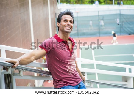 Man thinking of you. Wearing a red Polo shirt, blue jeans, stretching arms on the railing, a handsome, sexy, middle age guy is standing by a tennis court, looking up, smiling, thinking.