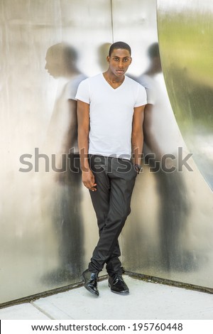 Street Fashion. Wearing a white V neck T shirt, pants, leather shoes, a hand in pocket,  a young black handsome man is standing in the corner of metal mirror walls, charmingly looking at you.