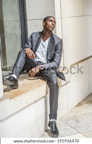 Young black man thinking outside. Wearing a white under wear, fashionable jacket, pants, shoes, a young black college student is sitting against a window frame, sad, emotionally looking up.