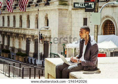 Young man working on street. A young black college student is sitting outside, working on a laptop computer, thinking.  Wall Street sign in the background.