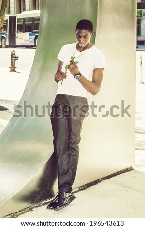 Teenager boy missing you.  Wearing a white V neck T shirt, pants, leather shoes, a young black guy is leaning back against  metal structure on street,  looking down at a white rose on hands, thinking.