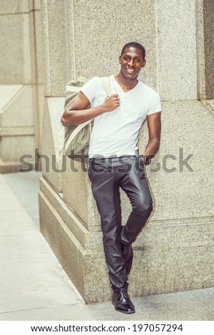 Street Fashion. Wearing a white V neck T shirt, pants, leather shoes, carrying a shoulder bag,  a young black college student is standing against a column outside, smilingly looking at you.