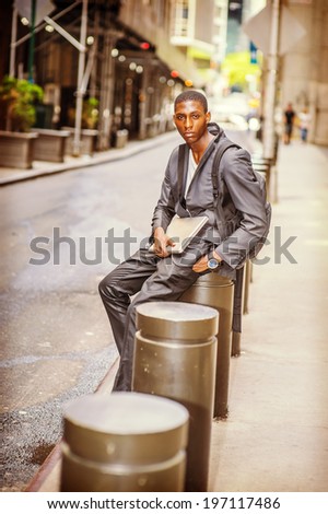 Young College Student. Carrying a bag, holding a laptop computer, a young handsome black guy is sitting on street, lost in thought.