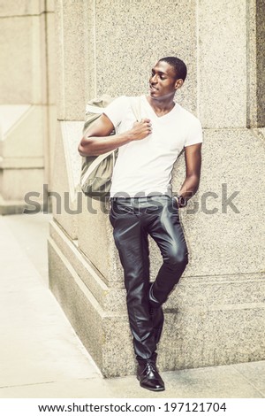 Street Fashion. Wearing a white V neck T shirt, pants, leather shoes, carrying a shoulder bag,  a young black college student is standing against a column outside, smilingly looking away.