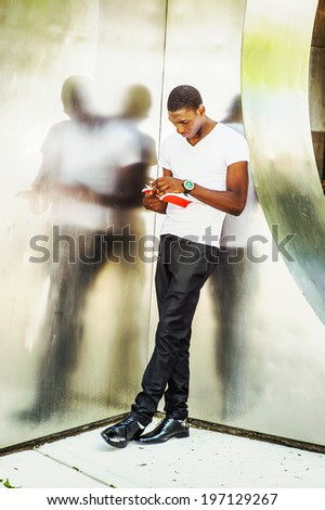Man Reading Outside. Wearing a white V neck T shirt, pants, leather shoes, wristwatch, a young black guy is leaning back against  metal mirror walls, looking down, turning pages, reading a red book.
