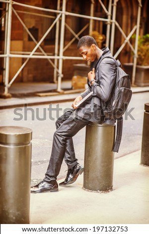 Young College Student. Carrying a bag, holding a laptop computer, a young handsome black guy is sitting on street, looking down, sad.