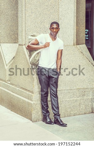 Street Fashion. Wearing a white V neck T shirt, pants, leather shoes, carrying a shoulder bag,  a young black college student is standing outside an office building, looking at you.