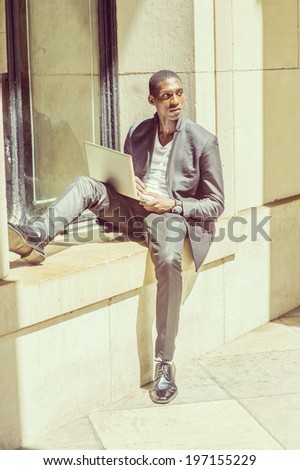 Young Man Studying Outside. Wearing a white under wear, fashionable jacket, pants, shoes, a young black college student is sitting against a window frame, working on a laptop computer.