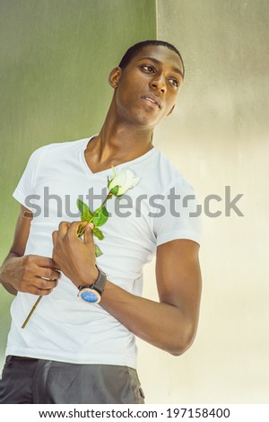 Young man missing you.  Wearing a white V neck T shirt, holding a white rose, a young black guy is leaning back against  metal structure, passionately looking up, lost in thought.