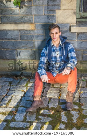 Man Casual Fashion. Wearing a blue and white pattern shirt,  a blue hood vest,  red jeans, brown leather boot shoes, a young guy is sitting on the ground against the wall with ivy leaves, relaxing.