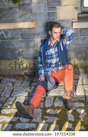 Man Casual Fashion. Dressing in blue, white pattern shirt, a hoody vest,  red jeans, leather boot shoes, holding a woolen Fedora hat on his knee, a young guy is sitting on the ground, thinking.