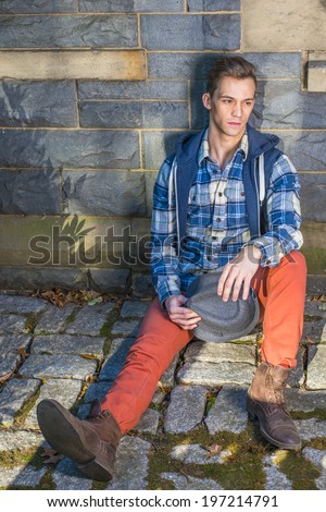 Man Casual Fashion. Dressing in blue, white pattern shirt, a hoody vest,  red jeans, leather boot shoes, holding a woolen Fedora hat, a young guy is sitting on the ground against wall, thinking.