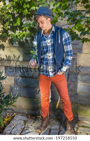 Man Casual Fashion. Dressing in blue, white pattern shirt, blue hoodie vest,  red jeans, brown leather boot shoes, wearing woolen Fedora hat, a young guy is holding white rose, looking down, thinking