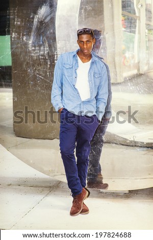 Man Casual Fashion. Wearing a white under wear, long sleeve shirt, blue pants, brown boot shoes, glasses on head, a young black guy is standing back against a mirror wall, casually looking at you.