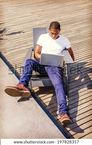 Man Working Outside. Wearing a white V neck T shirt, blue pants, brown boot shoes, a young black guy is casually sitting on a chair on wooden floor, looking down, working on a laptop computer.