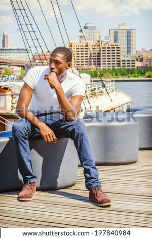 Man Thinking Outside. Wearing a white T shirt, blue pants, brown boot shoes, a young black guy is sitting on deck, a hand touching his mouth, relaxing, lost in thought. The background is a harbor.