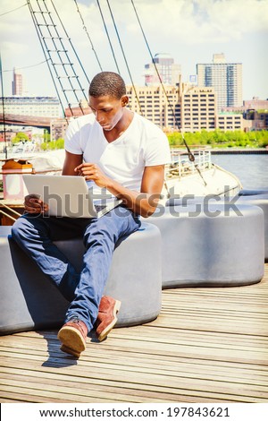 Man Working Outside. Wearing a white V neck T shirt, blue pants, brown boot shoes, a young black guy is sitting on deck, working on a laptop computer. The background is a harbor.