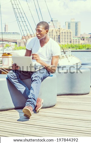Man Working Outside. Wearing a white V neck T shirt, blue pants, brown boot shoes, a young black guy is sitting on deck, working on a laptop computer, thinking. The background is a harbor.