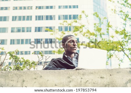 Young black man working outside. A young black college student is standing by the top of the wall outside an office building,  working on a computer.